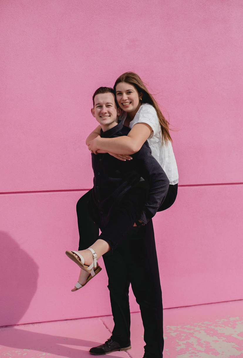 The Founders and Owners of Da Bored Book - Tyler & Kirsten standing with Kirsten holding Tyler in front of a pink wall